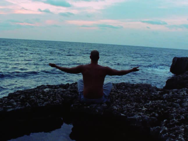 A shirtless man doing yoga on a rock formation next to the ocean