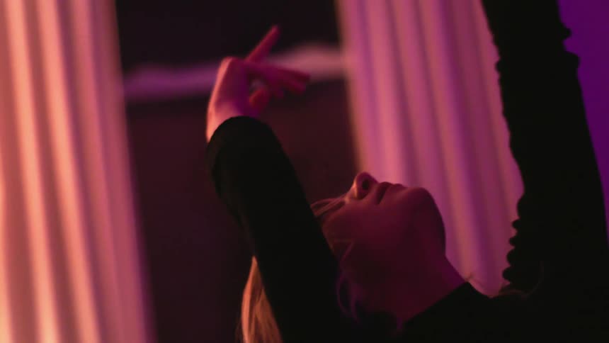 Blonde woman with black sweater dances slowly as lights flash