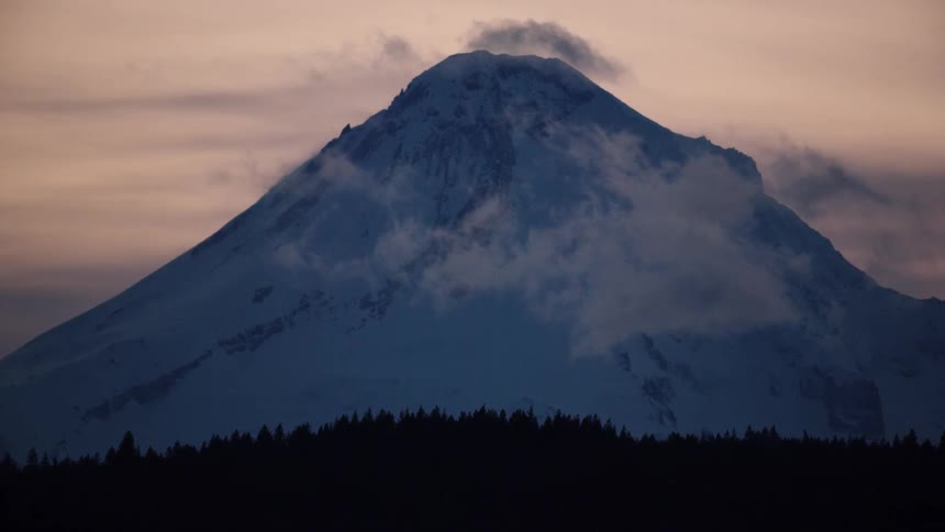 High winds blow clouds rapidly past the summit of Mount Hood after sunset with silhouetted ridges on the bottom of the frame
