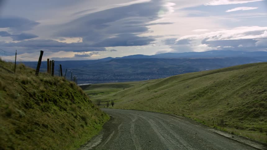 Lenticular clouds fill the sky in dynamic formations as the vehicle drives up a twisty and winding dirt road with green meadows and old barb wire fences on both sides