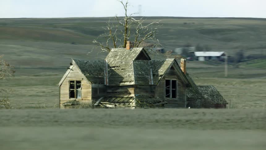 An old farmhouse decays in a rural field as the vehicle passes by from the perspective of a passing vehicle