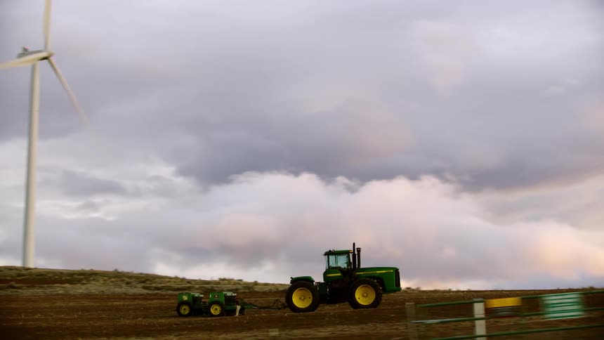 The camera follows a parked John Deere tractor at the base of several wind turbines on a wind farm with beautiful sunset clouds in the background