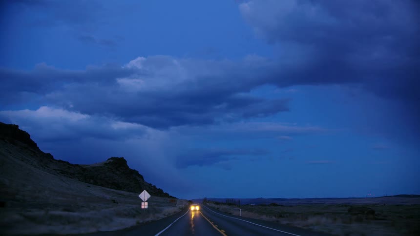 A lone driver works their way home after a long day on the ranch as they navigate a lone two lane highway after sunset with blue puffy clouds in the sky