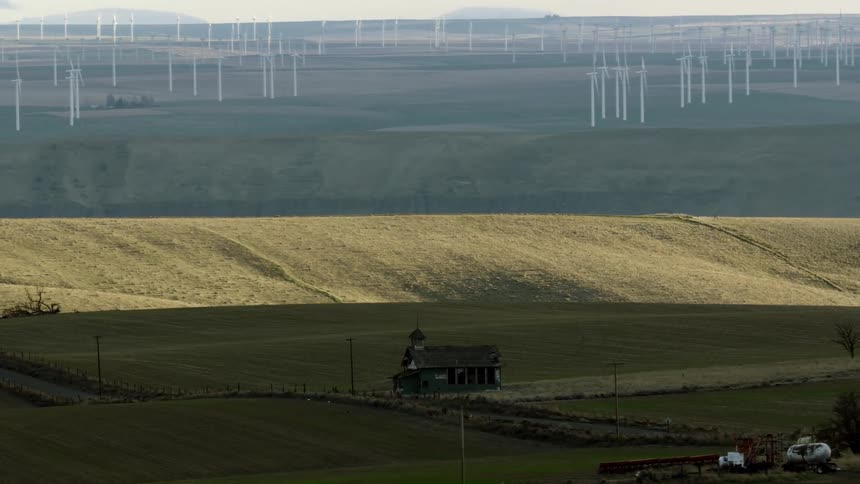 An old school house sitting on the hill with a large wind farm beyond it on the horizon as the wind mills are rotating creating renewable clean electricity for the USA