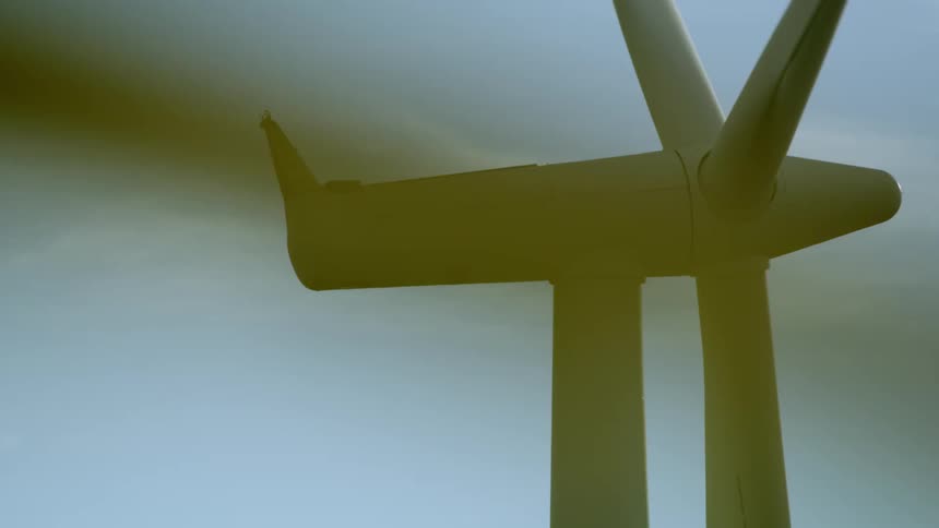 The blades on a wind turbine spin in the wind in this tight shot following the turbine before it is obscured by a ridge in a natural wipe transition