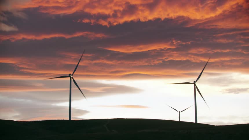 Unbelievable sunset shot of three silhouetted wind turbines rotating with energy as the clouds erupt into orange, purple and pink colors showing the beauty of renewable energy