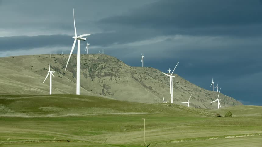 Wind turbines rotate as afternoon sunlight illuminates them brightly contrasting the dark storm clouds far beyond the large mountain in the background