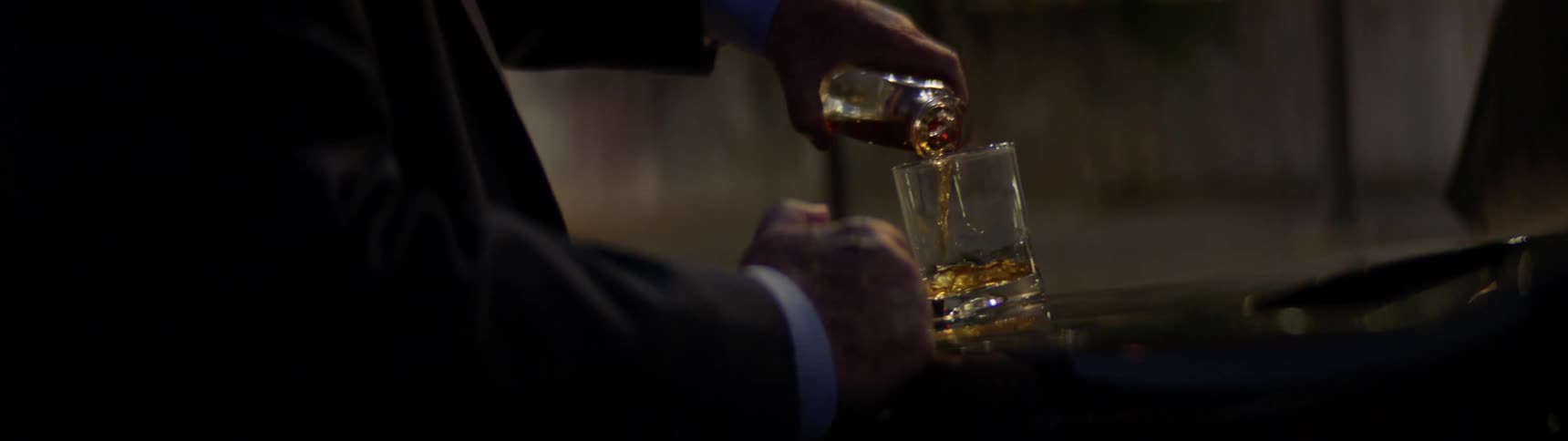 An older white man in a suit pours a drink on the hood of his car