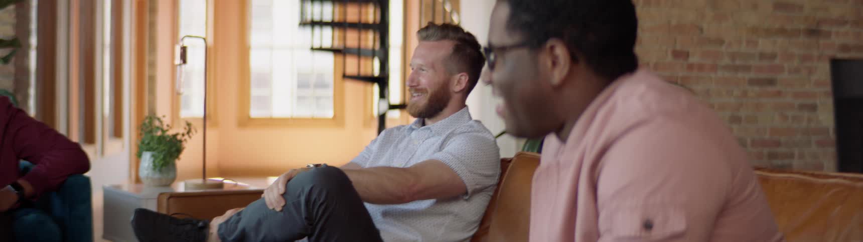 A black man and a white man sit in a living room on a couch laughing and having a good time