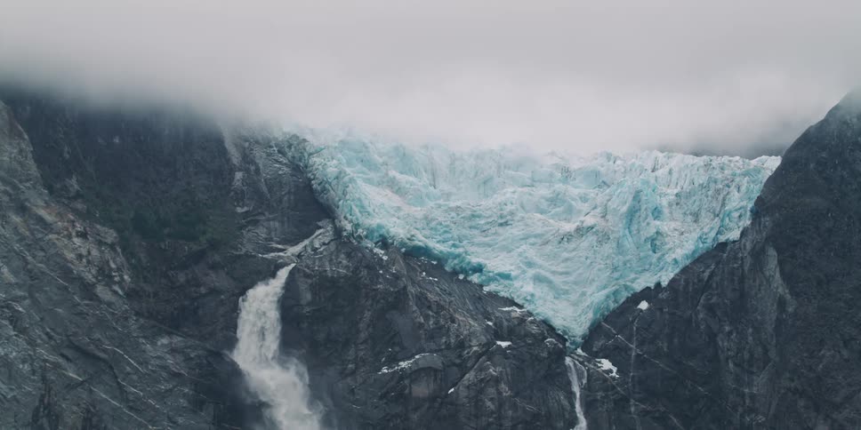 A glacier sits atop a rocky mountain with waterfalls flowing from underneath it