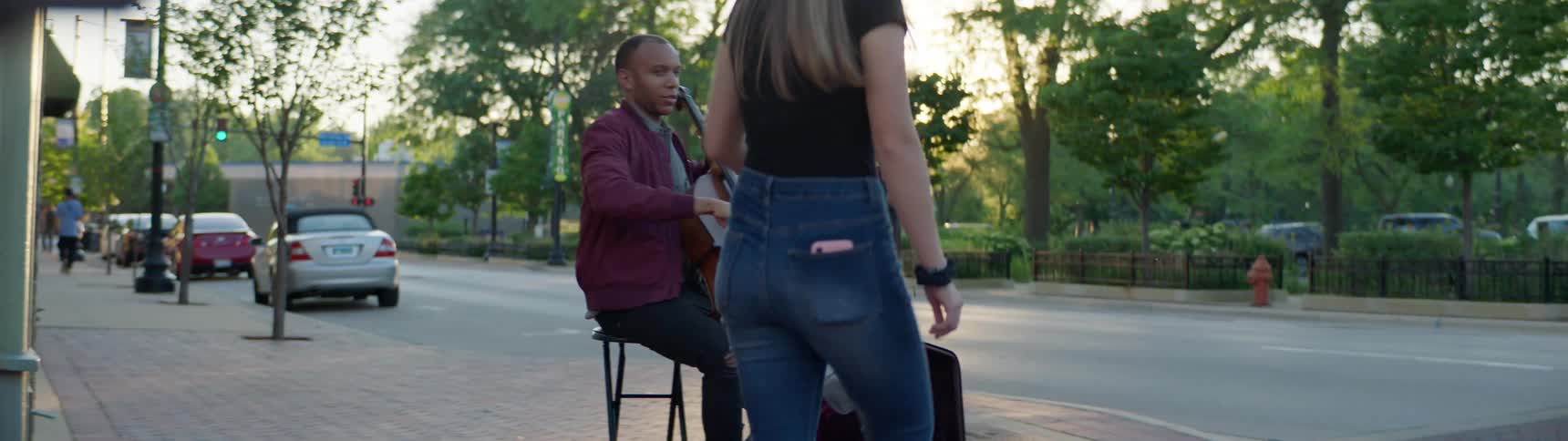 Two white women give money to a black man busking with a cello on a street corner