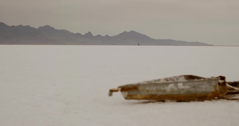 A person walks across the desert as a destroyed boat sits on the sand