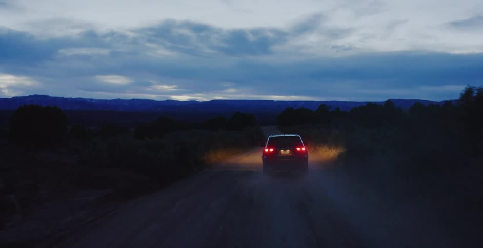 Car with headlights illuminating the road drives on dirt road at dusk in the country