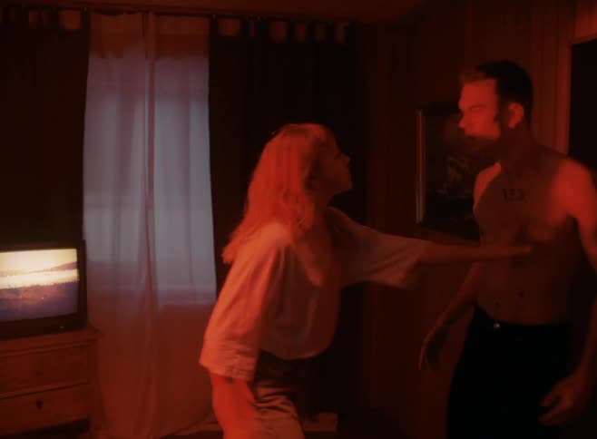 Shirtless man and woman with blonde hair in red light argue with each other in a room