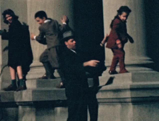 A father wearing a suit lifts his daughter up and places her on a pedestal of a classical style building