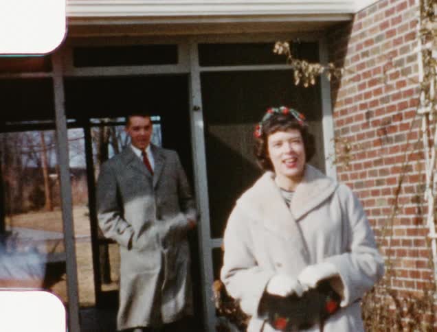 A man comes out of a house wearing a coat and tie and reaches for a woman who is talking as she walks down the steps