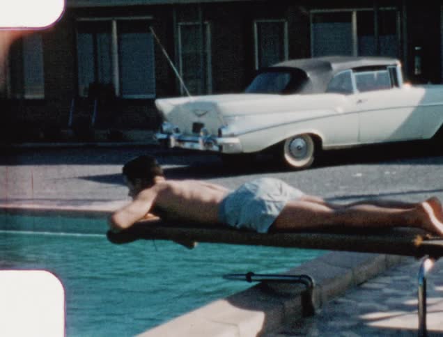 A man lies on a diving board and puts his hand in the swimming pool below
