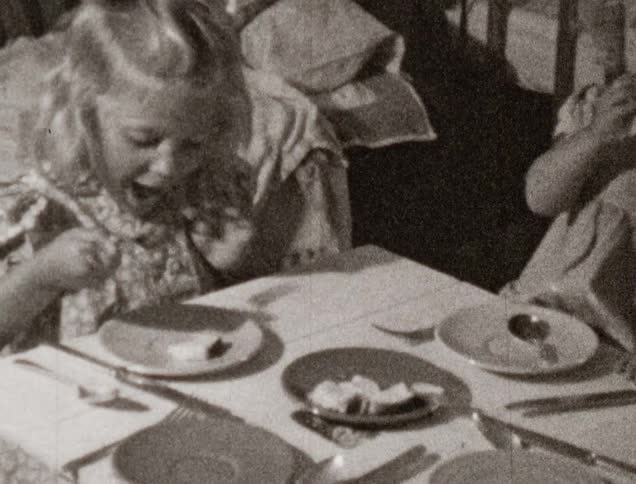 Two little girls eat cake at a small kids table covered in a cloth and set for four