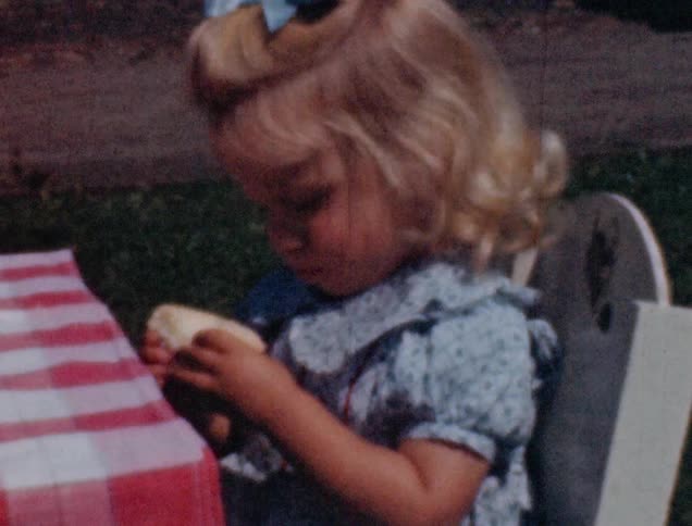 A little blonde girl in a blue dress sits at a table outside and eats a piece of cake