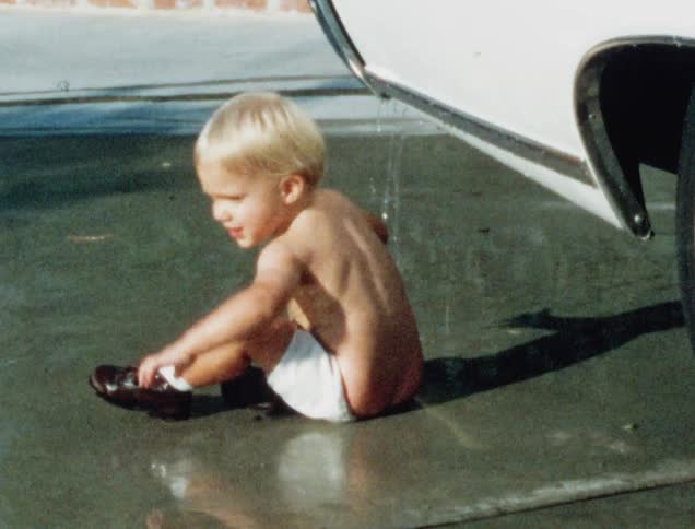 A white male toddler touches water dripping off of a car