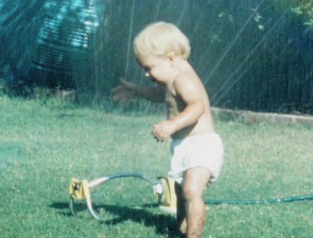 A toddler wearing a diaper touches water coming out of a sprinkler then runs away