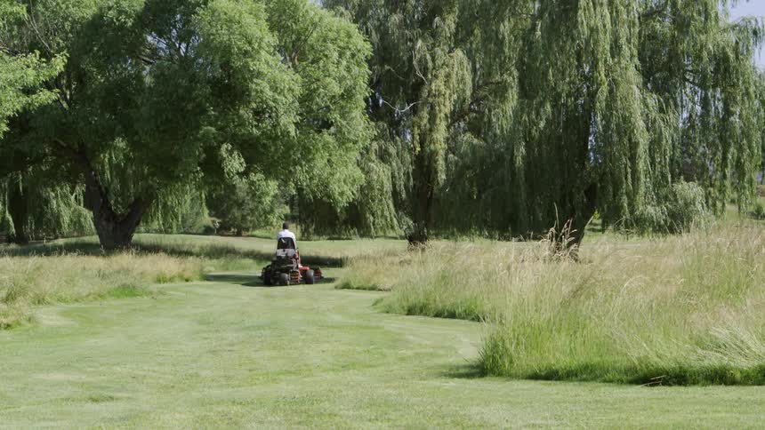 A man steers a riding lawn mower over part of a golf course