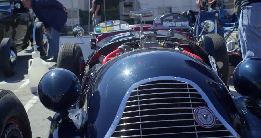 A racing car sits in a parking lot surrounded by white men and other cars