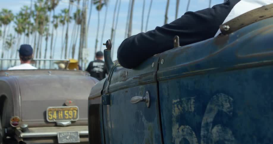 Vintage vehicles lined up on a street with their drivers sitting inside wearing old fashioned clothing