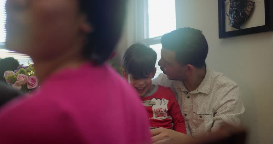 A man holds his son on his lap sitting in a chair by a window