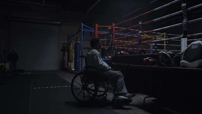 A man in a wheelchair sits beside a boxing ring in the dark