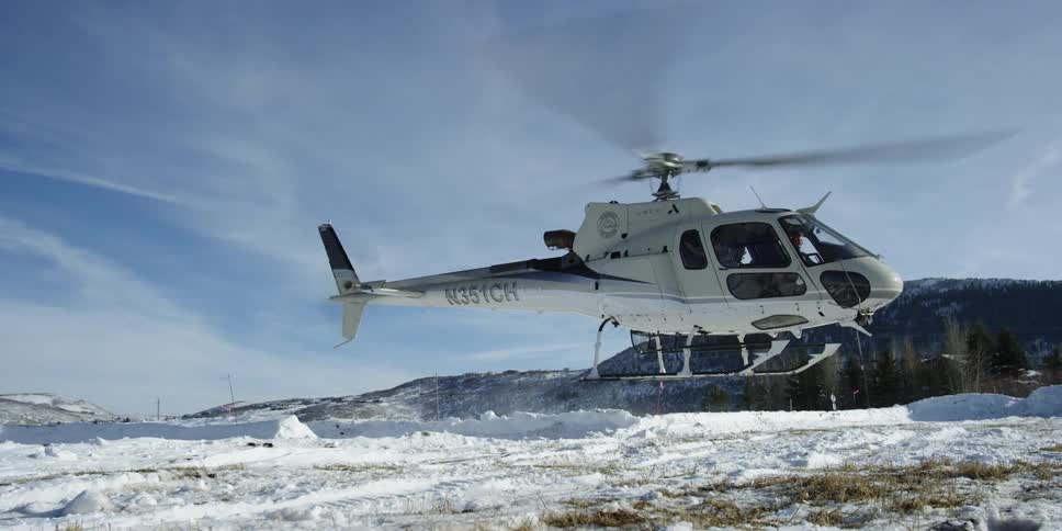 A helicopter rises kicking up snow and blowing grass on a bright sunny day