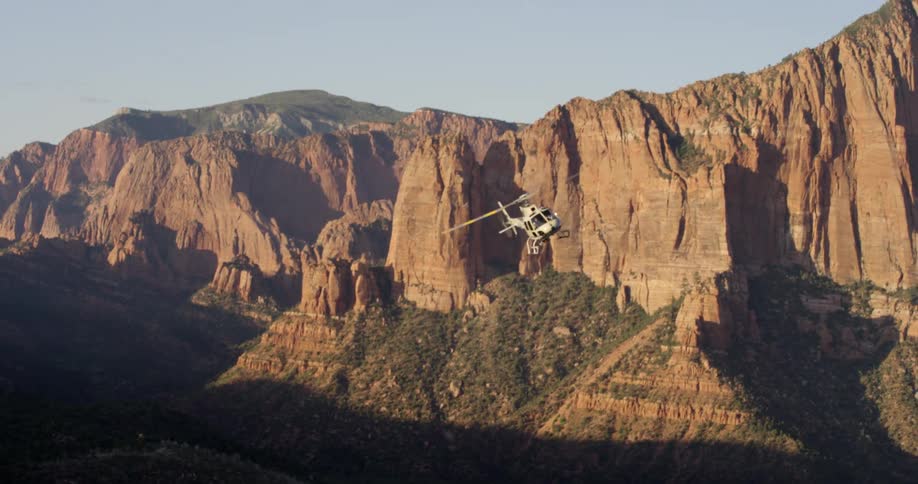 A helicopter rises out of a canyon near a rocky ridge and cliff and flies off