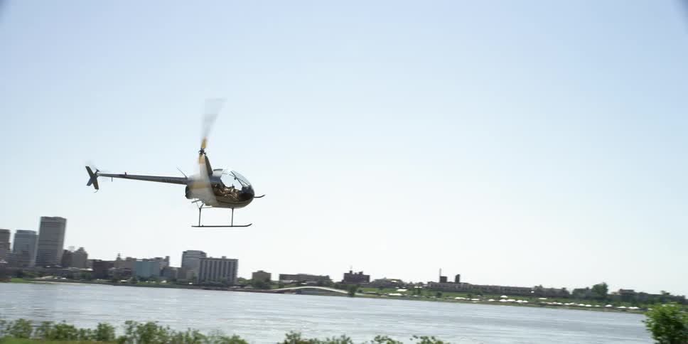 Helicopter flies over a river that runs between city buildings and a park