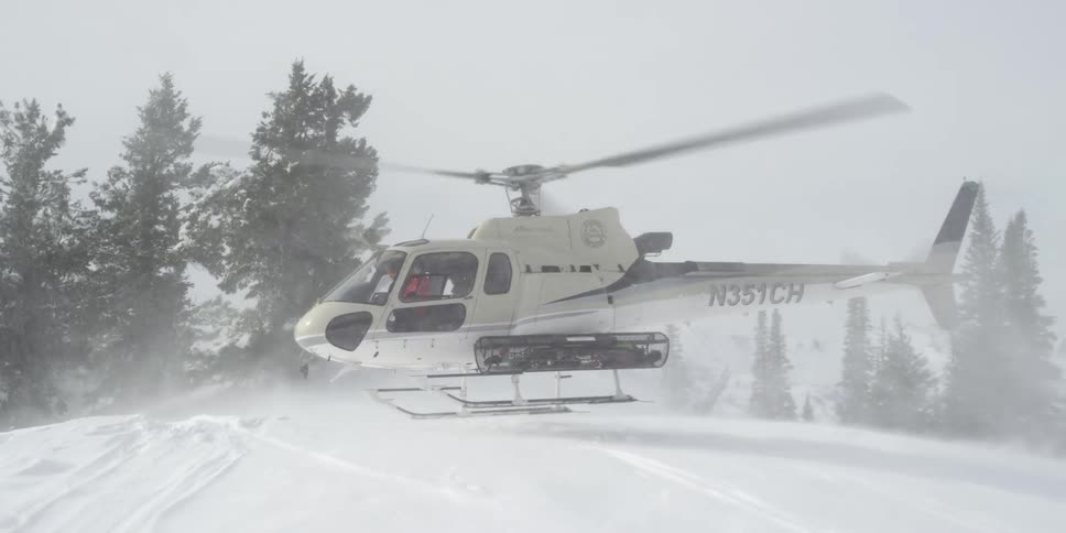 White helicopter hovers over snow covered slope near conifers blowing snow into air