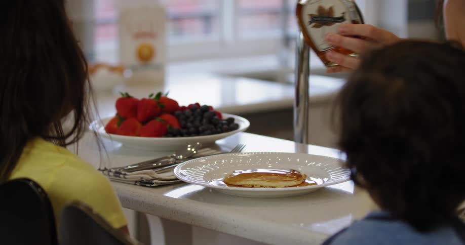 Daughter and son sitting at kitchen counter watch as their mother pours maple syrup onto a plate of pancakes