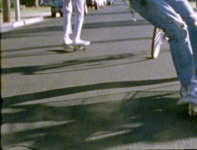 A group of friends push another in a shopping cart and ride skateboards and bicycles on a neighborhood street