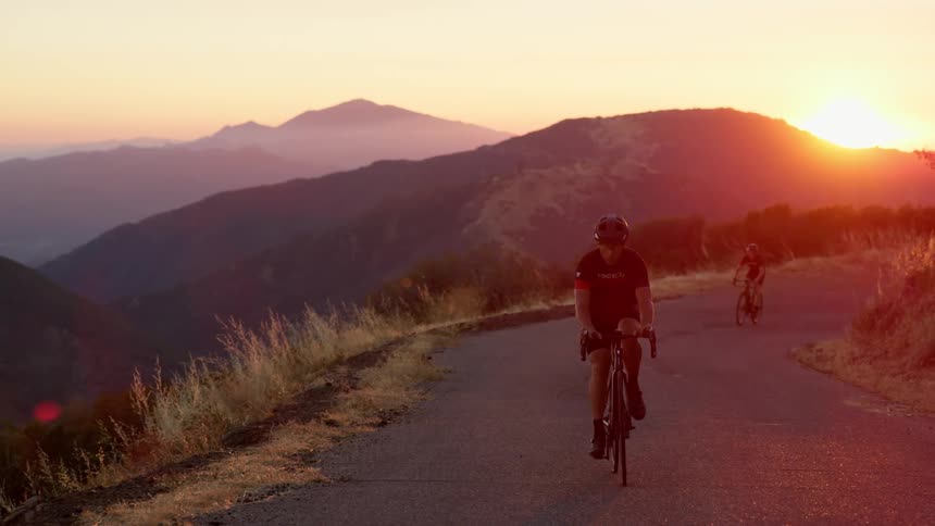 Two people riding bikes on a road on a cliff with the sky and mountains in the background
