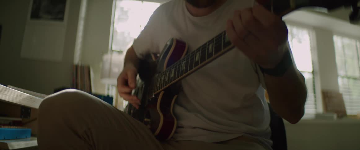 Man with a white t shirt and black headphones plays a electric guitar as the sunlight comes through the window