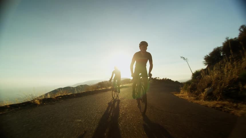 Two men riding bikes on a road on a cliff with the sky and mountains in the background