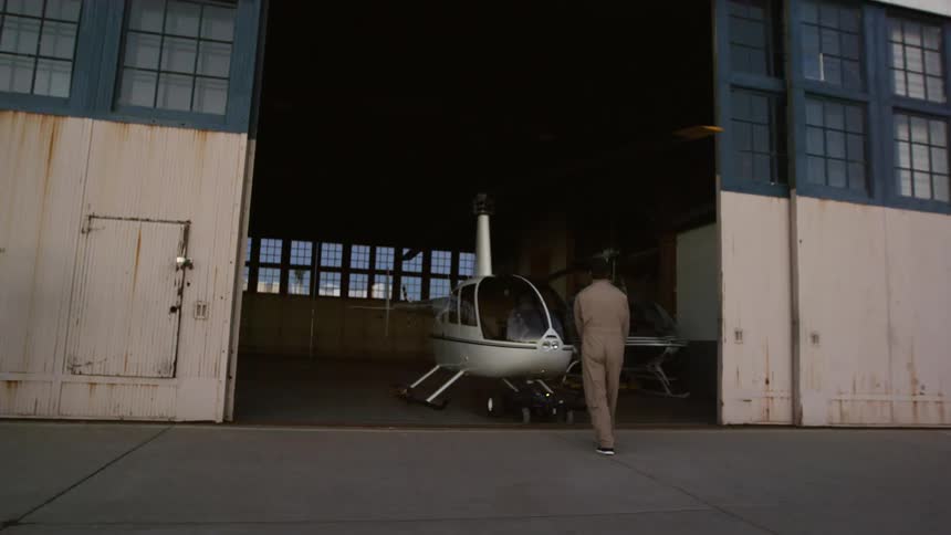 A man looks on as a worker moves a small helicopter into a hangar with a hand cart