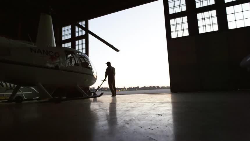 A man walks to the front of a helicopter in a hangar and grips the handle of a helicopter tow cart