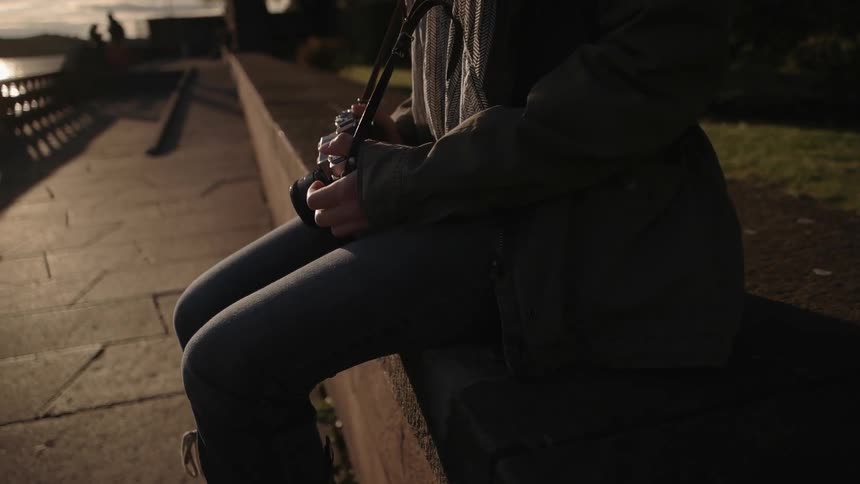 A female photographer sitting on a knee wall next to a body of water at sunset