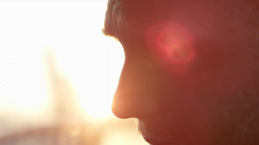 Man withstubble beard sits in a car moving across the road at sunset