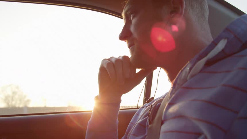 A white man sits in the back seat of a car while wearing ear buds