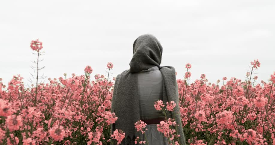 A woman stands in a field of pink flowers with her head and hair wrapped in a gray scarf on a cloudy day