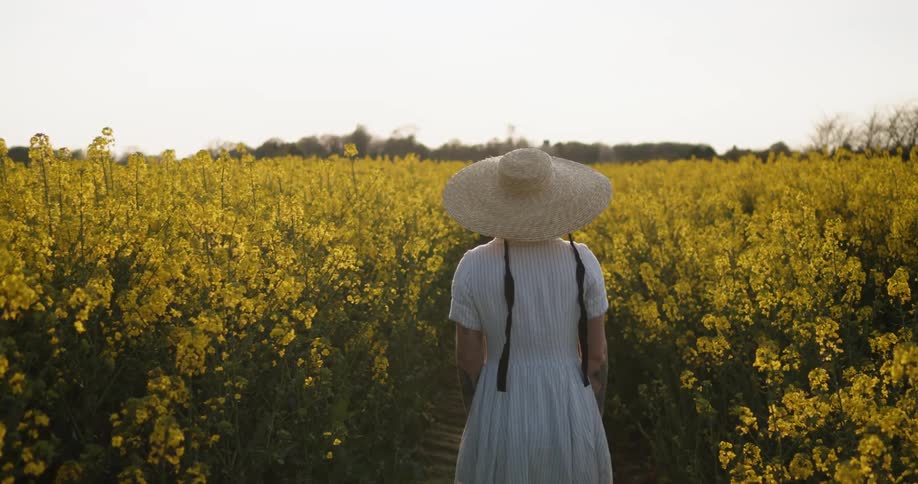 Woman wearing large sun hat walking through a field of yellow flowers in the country