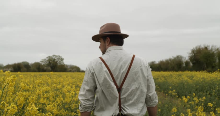 Man wearing suspenders and a hat walks through a field of yellow wildflowers