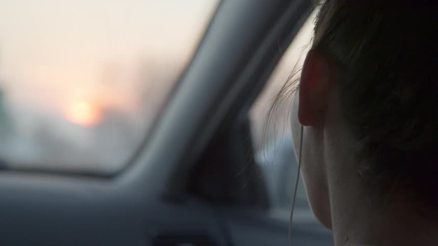 A young woman rides in a vehicle at dusk while wearing earbuds