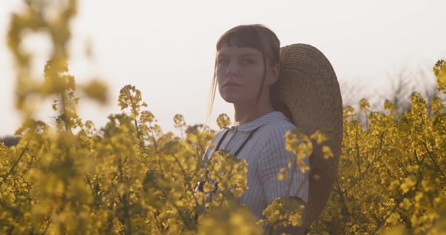 A woman in a dress stands alone among tall yellow flowering plants in a field