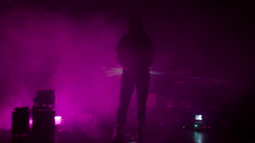 A white woman stands amidst strobe lights in a room with machines stacked on the floor
